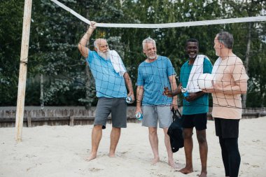 smiling multiracial elderly men with volleyball on sandy beach clipart