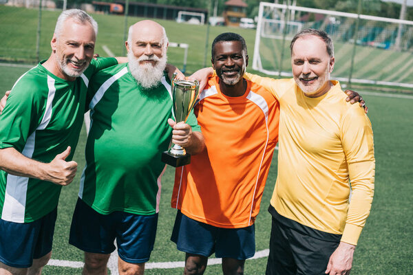 многонациональные улыбающиеся старые спортсмены с кубком чемпионов, стоящим на футбольном поле
