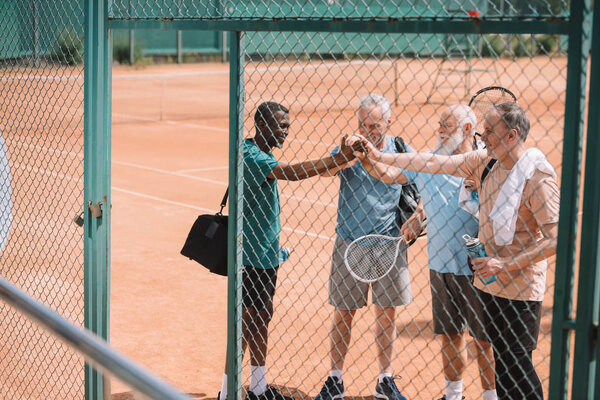 multicultural group of elderly tennis players holding hands together after game on court