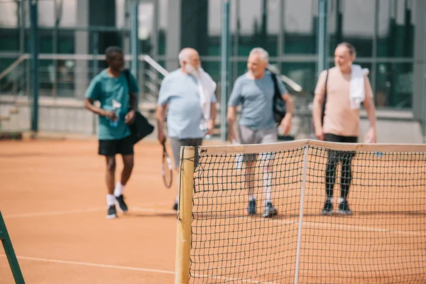 selective focus of multiracial elderly friends with tennis equipment on court