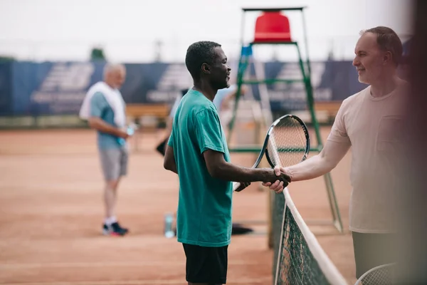 selective focus of interracial elderly friends with tennis racquets shaking hands after game on court