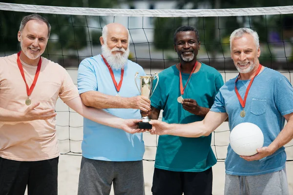 portrait of smiling multiethnic elderly friends with tennis ball, medals and champions cup on beach