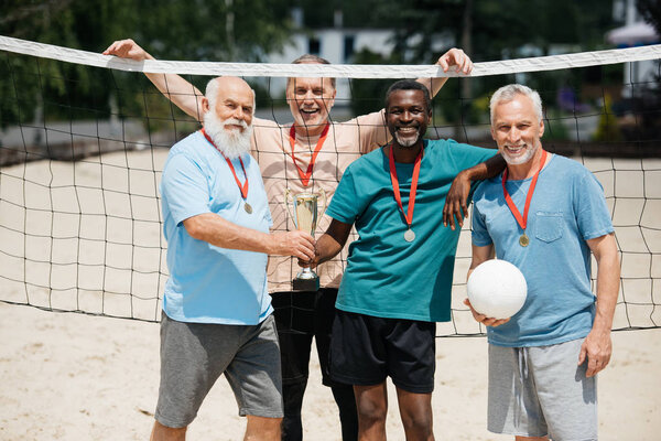 портрет улыбающихся многонациональных пожилых друзей с теннисным мячом, медалями и кубками чемпионов на пляже
