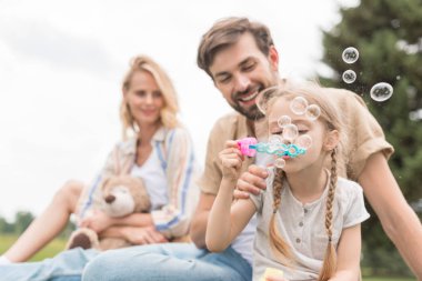 oyuncak ayı tutarak ve baba ve kız parkta sabun köpüğü üfleme bakarak anne