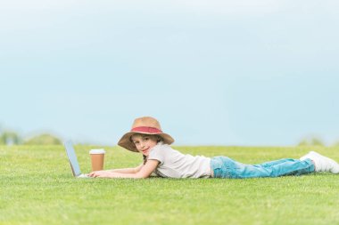 şapkalı yeşil çayır on laptop kullanırken kameraya gülümseyen sevimli çocuk