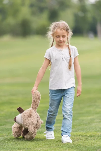 テディベアを押しながら緑の芝生の上を歩く愛らしい小さな子供  — 無料ストックフォト