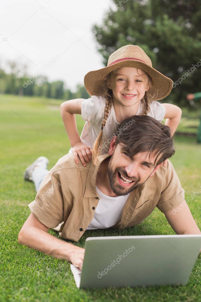 happy father and daughter using laptop and smiling at camera in park 