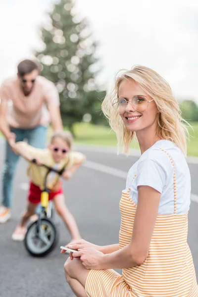 幸せな若い女スマート フォンを押しながら後ろに自転車に乗る父教え娘ながらカメラに笑顔 — ストック写真