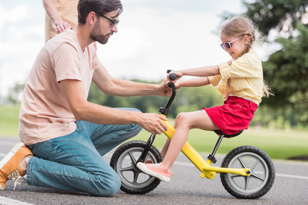 вид сбоку на молодого отца, обучающего маленькую дочь езде на велосипеде в парке
