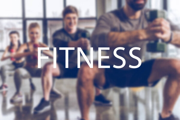 blurred group of athletic young people in sportswear with dumbbells exercising at gym, fitness inscription