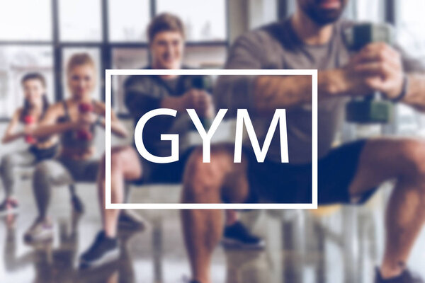 blurred group of athletic young people in sportswear with dumbbells exercising at gym, gym inscription