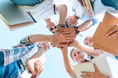 bottom view of happy multiethnic teenage classmates stacking hands in park  