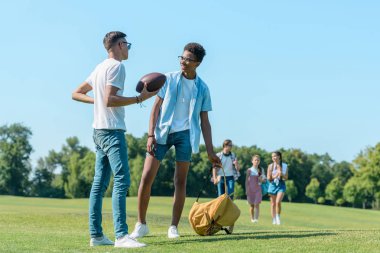 multiethnic boys playing with rugby ball while classmates walking behind in park  clipart