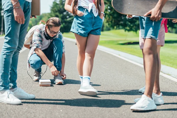 青少年用书本和滑板在公园里等待朋友绑鞋带的镜头 — 免费的图库照片