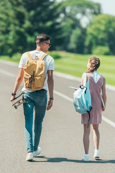 十代の女の子とバックパック 歩いて 公園で一緒に話してはスケート ボードを持つ少年の背面図 — ストック写真