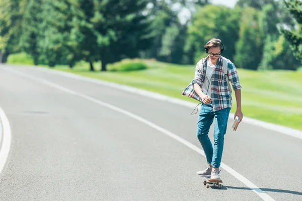 teenage boy in headphones holding books and riding skateboard in park