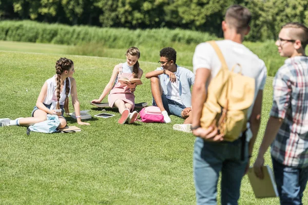 Estudantes Adolescentes Multiétnicos Sentados Grama Estudando Enquanto Colegas Andando Primeiro — Fotografia de Stock