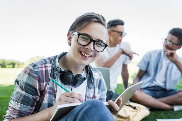 Feliz Adolescente Tomando Notas Sonriendo Cámara Mientras Estudia Con Amigos — Foto de Stock