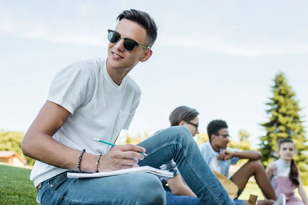 Smiling Teenage Boy Sunglasses Taking Notes Looking Away While Studying — Stock Photo, Image