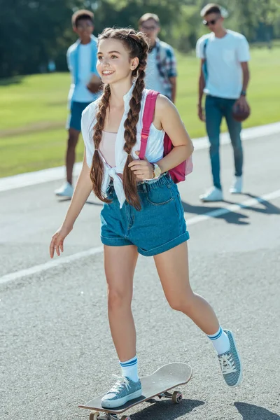微笑的少女与背包骑滑板 而同学在公园里走在后面 — 图库照片