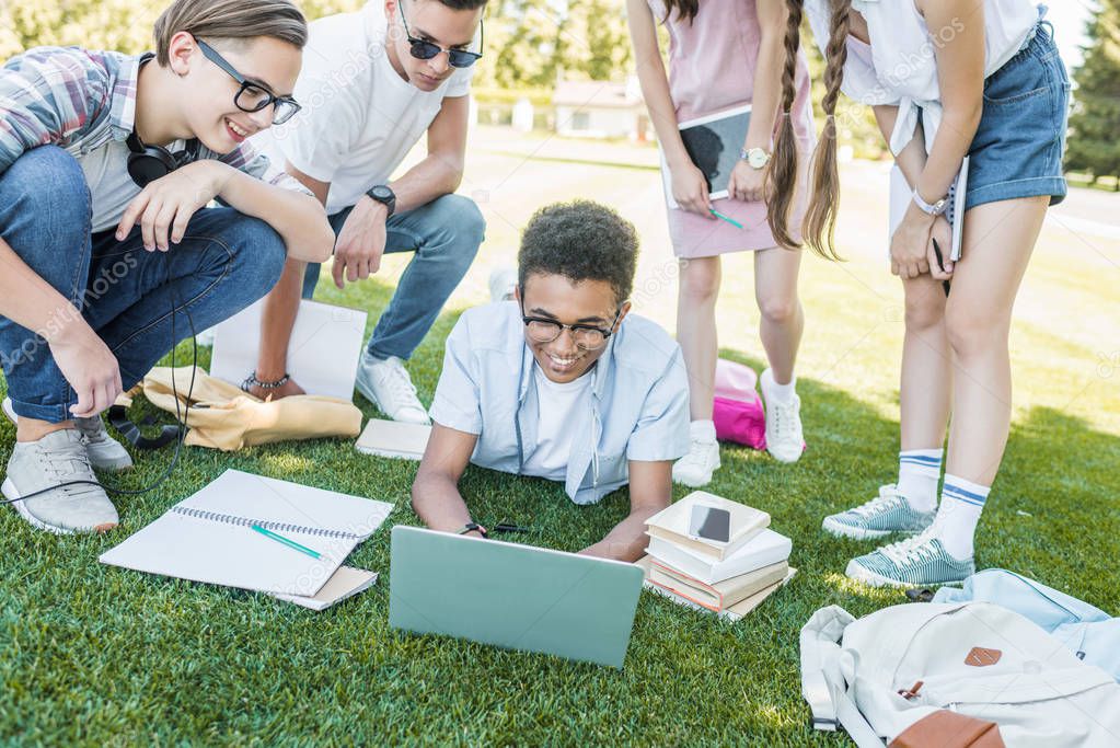 happy multiethnic teenage students studying with books and digital devices in park