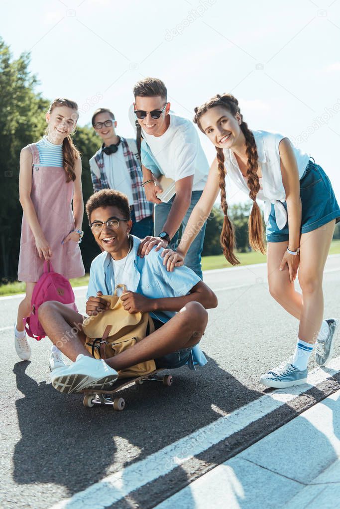 cheerful teenage multiethnic classmates with books and backpacks smiling at camera while having fun with skateboard in park