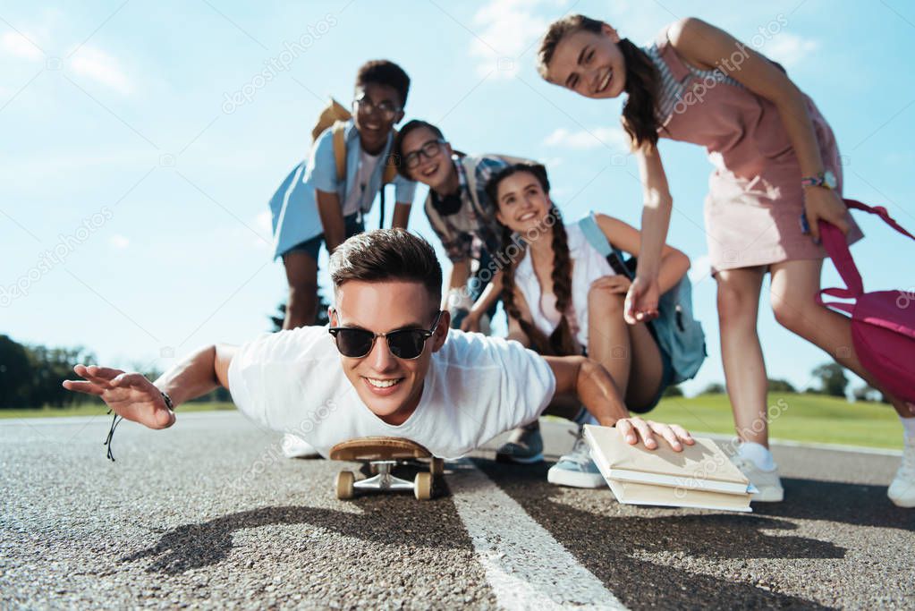 happy teenage multiethnic friends looking at smiling boy lying on skateboard in park