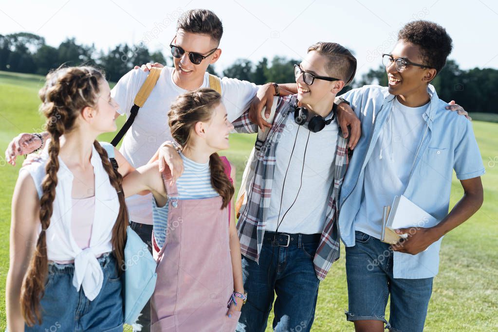 happy multiethnic teenagers holding books and smiling each other in park