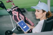 Seitenansicht einer Golfspielerin mit Tablet mit Facebook-Logo auf dem Bildschirm in den Händen auf dem Golfplatz