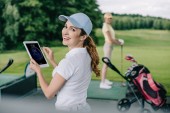 Selektiver Fokus einer lächelnden Frau mit Tablet und Freundin beim Golfspielen auf dem Golfplatz