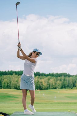 side view of woman in polo, sunglasses and cap playing golf at golf course clipart