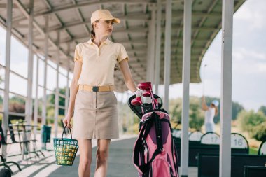 portrait of woman in polo and cap with golf gear walking at golf course clipart
