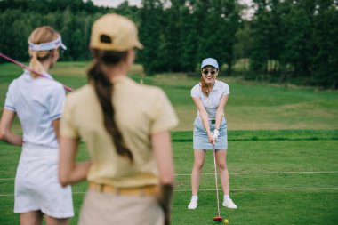 selective focus of women in caps with golf equipment looking at friend playing golf at golf course clipart