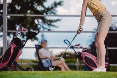 kadın golf oyuncu oynarken golf golf sahasında arkasında oturan arkadaşı ise kısmi görünümünü
