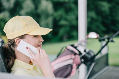 smartphone üzerinde golf sahasında konuşurken şapkalı kadın golf oyuncu yan görünüm