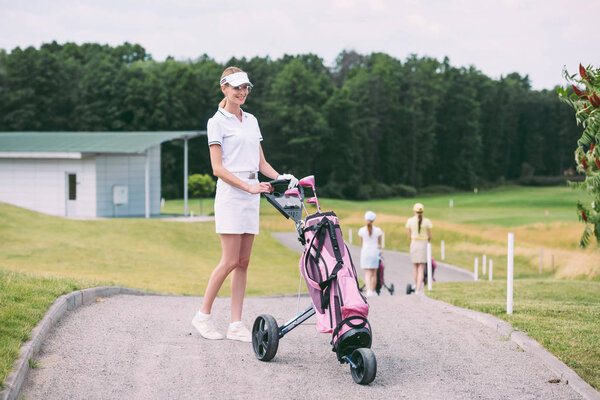 selective focus of cheerful female golf player in cap and white polo with golf gear at golf course