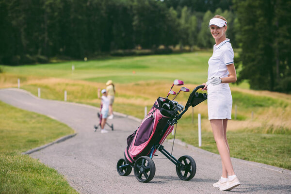 selective focus of smiling female golf player in cap and white polo with golf gear at golf course