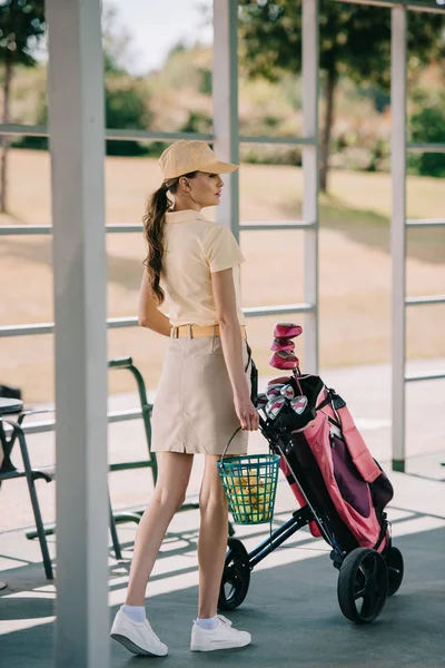 side view of attractive woman in polo and cap carrying golf equipment at golf course