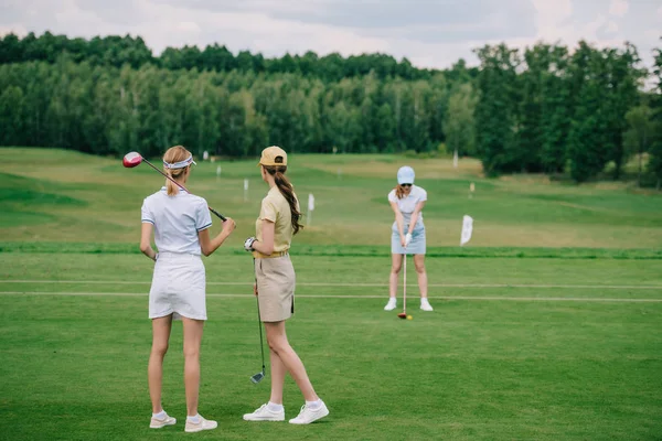 selective focus of women in caps with golf equipment looking at friend playing golf at golf course