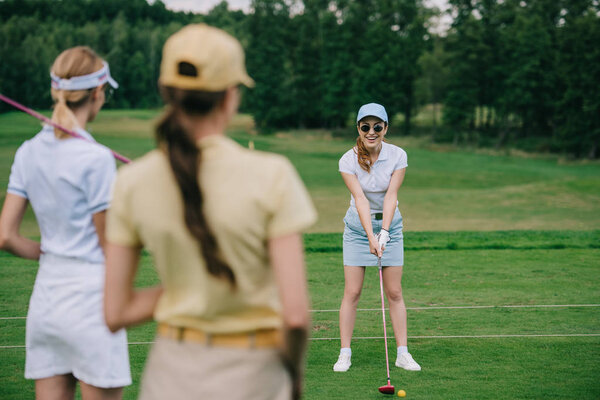 selective focus of women in caps with golf equipment looking at friend playing golf at golf course