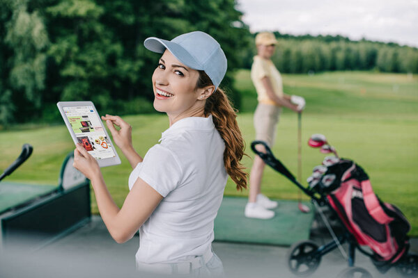 selective focus of smiling woman holding tablet with ebay logo and friend playing golf behind at golf course