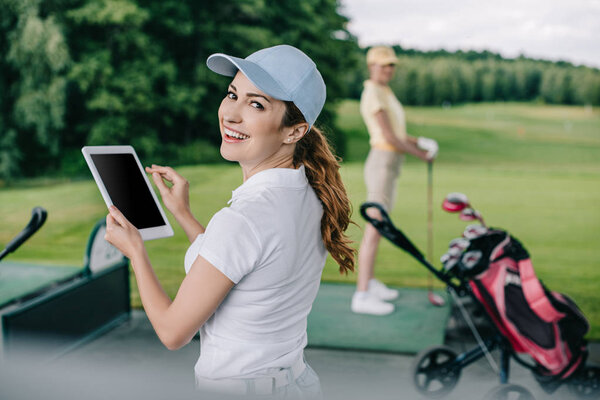 селективный фокус улыбающейся женщины с планшетом и другом, играющей в гольф позади на поле для гольфа
