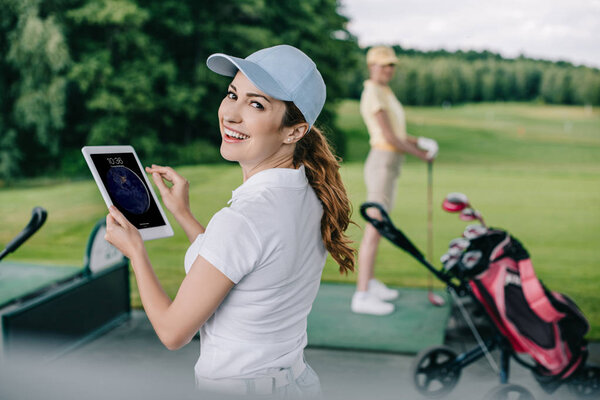 selective focus of smiling woman with tablet and friend playing golf behind at golf course