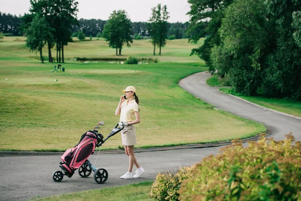 ゴルフギア ゴルフ コースを歩きながらスマート フォンで話している女性のゴルフ プレーヤーの側面図  — 無料ストックフォト