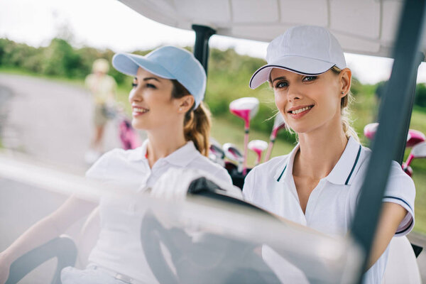 portrait of smiling female golfers in caps riding golf cart at golf course