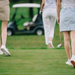 Cropped shot of women with golf clubs walking on green lawn at golf course