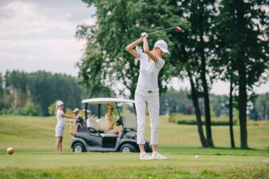 selective focus of woman with golf club playing golf and friends resting at golf cart on green lawn clipart