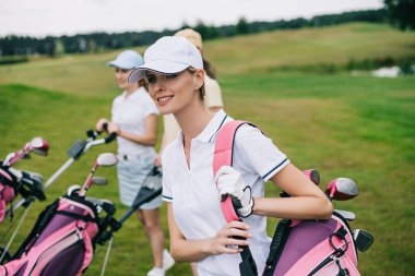 smiling women in caps with golf equipment on golf course clipart