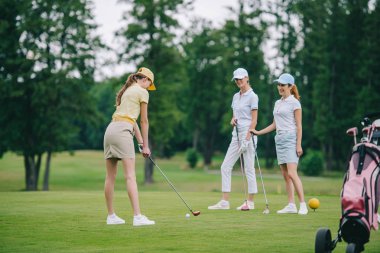 woman in cap playing golf while smiling friends standing near by at golf course clipart