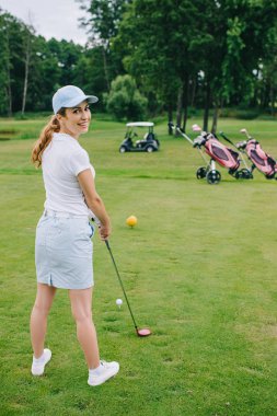 side view of smiling female golf player in cap with golf club standing at golf course clipart